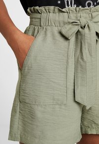Světlé zelené šortky z texturované látky, s vysokým pasem, zavazováním v pase a velkými bočními kapsami. Okraje jsou oškubané pro zajímavý detail.