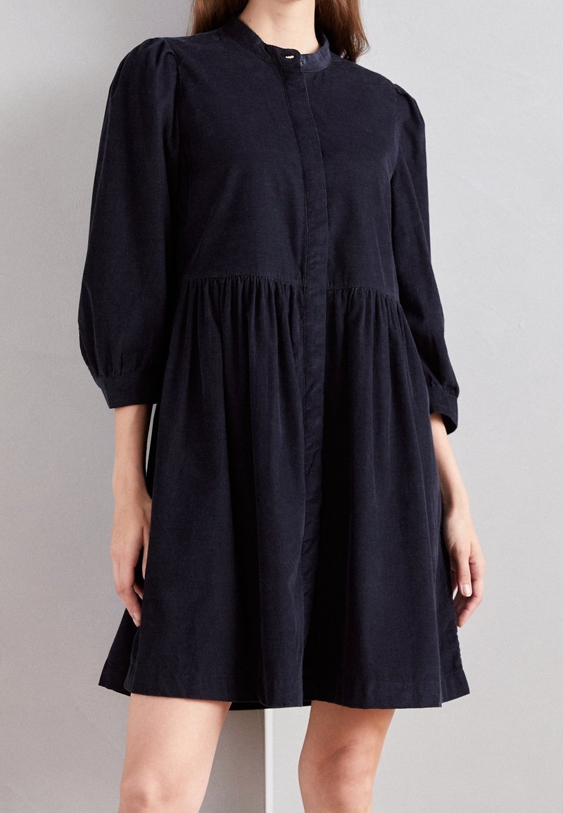 Femme portant une robe noire à manches longues boutonnée, avec une taille froncée et un ourlet court, sur un fond gris uni.