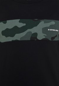 Camiseta negra con un patrón de camuflaje verde en el pecho, textura suave y la marca "G-STAR RAW" en blanco en la parte inferior derecha.
