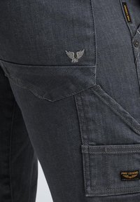 Dunkelblaue Jeans haben eine strukturierte Oberfläche, ein gesticktes Logo, Fronttaschen und eine auffällige Klapptasche mit einem Etikett an der Seite.