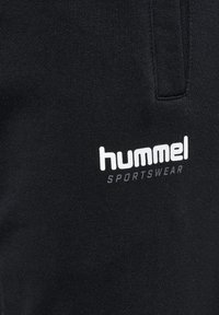 Schwarze Sportbekleidungshosen mit geripptem Material, elastischem Taillenbund und einem weißen "hummel"-Logo mit "SPORTSWEAR" darunter.