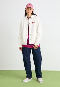 Jachetă din denim alb cu cusături roșii și logo, purtată peste un pulover roz cu gradient și blugi negri, completată cu pantofi bej și o șapcă roz.