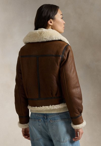 Woman wearing brown shearling jacket with cream fur collar and cuffs, paired with blue denim jeans, shown from back and side profile.