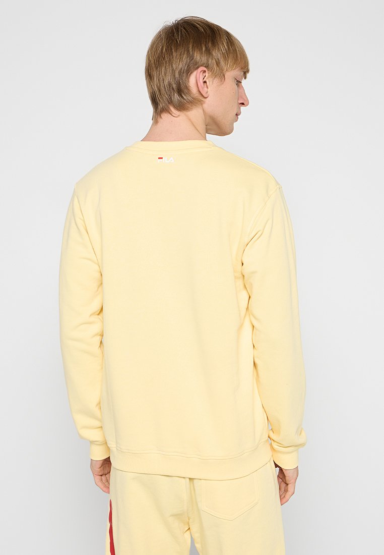 Sudadera de felpa amarilla con mangas largas, puños y dobladillo acanalados, que presenta un pequeño logotipo en el cuello, textura suave y corte relajado.