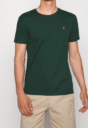 Hombre que lleva una camiseta de manga corta color verde oscuro con un pequeño logo bordado en el pecho y pantalones cortos beige sobre fondo liso.