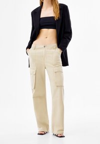 Beige cargobyxor med en avslappnad passform, flera sidofickor och knäppning med knapp. Tillverkade av ett slätt, hållbart tyg. Styla med en svart topp.