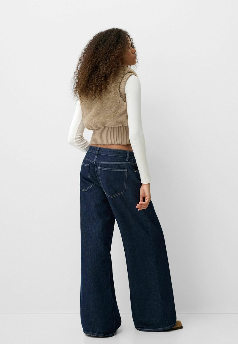 Beige Kunstfell-Cropped-Weste über einem weißen Langarmoberteil, kombiniert mit dunkelblauen, weit geschnittenen Jeans. Mit gerippten Akzenten und detaillierter Stickerei.