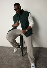Dunkelgrüne gepolsterte Weste, weißes Langarmshirt, hellgraue Jogginghose mit Seitenstreifen und weiße Turnschuhe in sitzender Pose auf einem schwarzen Hocker.