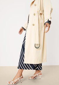 Beige trench coat with large buttons, cinched waist, and a green accent on the belt. Worn over a navy and white striped dress and white heeled sandals.
