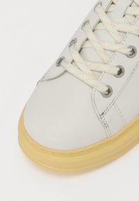 Weiße Ledersneaker mit runder Zehenpartie, gelber Gummisohle und metallischen Ösen. Mit weißen Schnürsenkeln und gelben Akzenten.