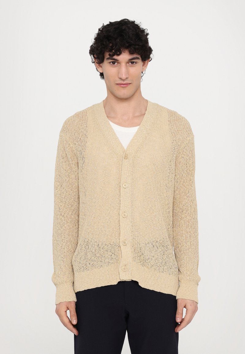 Beige strikket cardigan med V-hals, frontknapper og ribbestrikkede mansjetter. Teksturen er løst strikket, noe som gir et lett utseende.