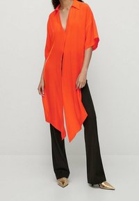 Blouse orange fluide avec un décolleté profond et des manches courtes, associée à un pantalon noir à jambes larges et des ballerines pointues dorées.