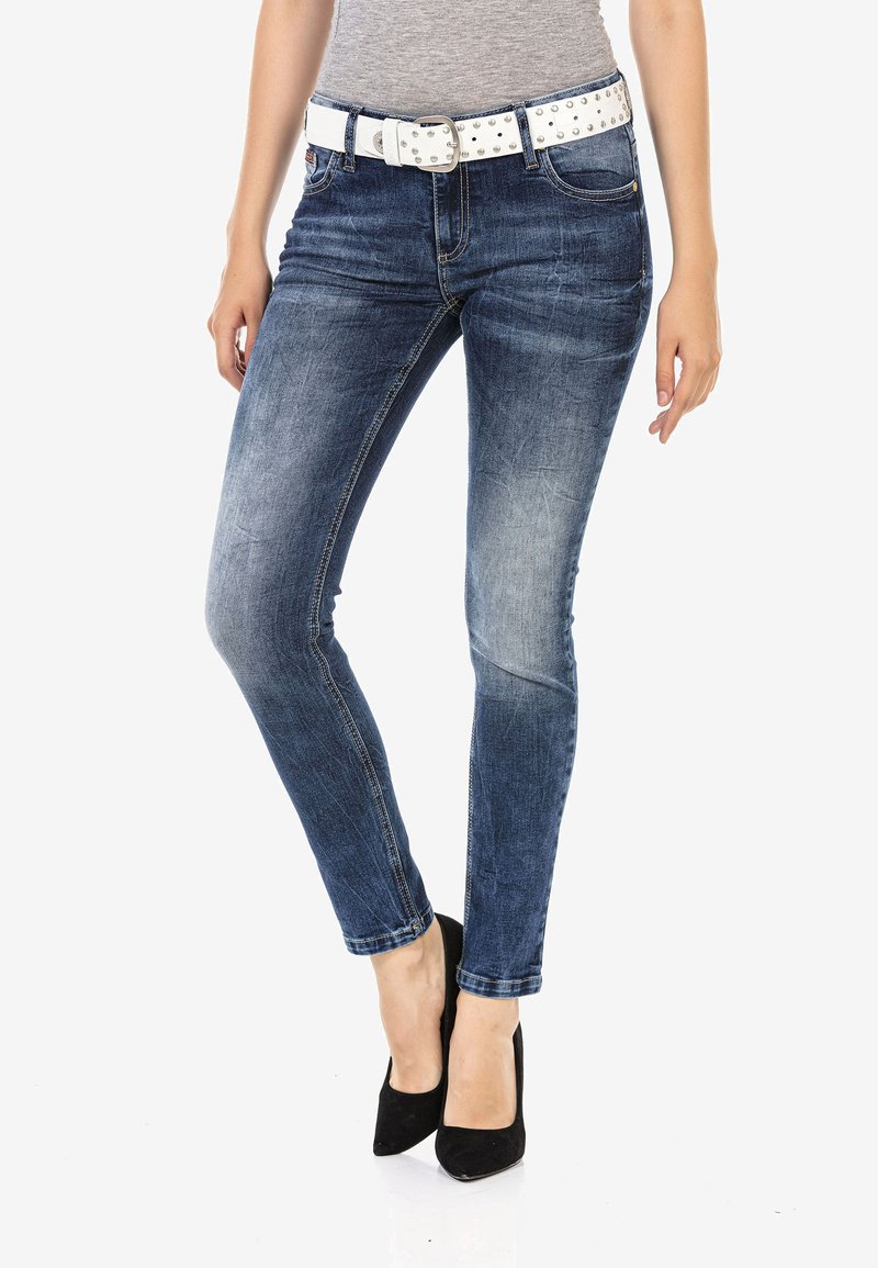 Jeans en denim slim de couleur bleu foncé avec un léger délavage, assortis à une ceinture blanche cloutée. Le mannequin porte des escarpins noirs à bout pointu.