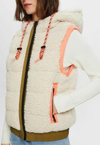 Fleece gevoerde bodywarmer met capuchon, crèmekleurig, oranje accenten, zwarte ritssluiting, voorzakken met roze bies en geribbelde witte lange mouwen.