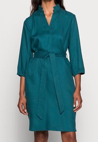 Teglrød kjole lavet af blødt stof med flæsekant ved kraven, 3/4-ærmer og et knyttet bælte i taljen for en markeret silhuet.