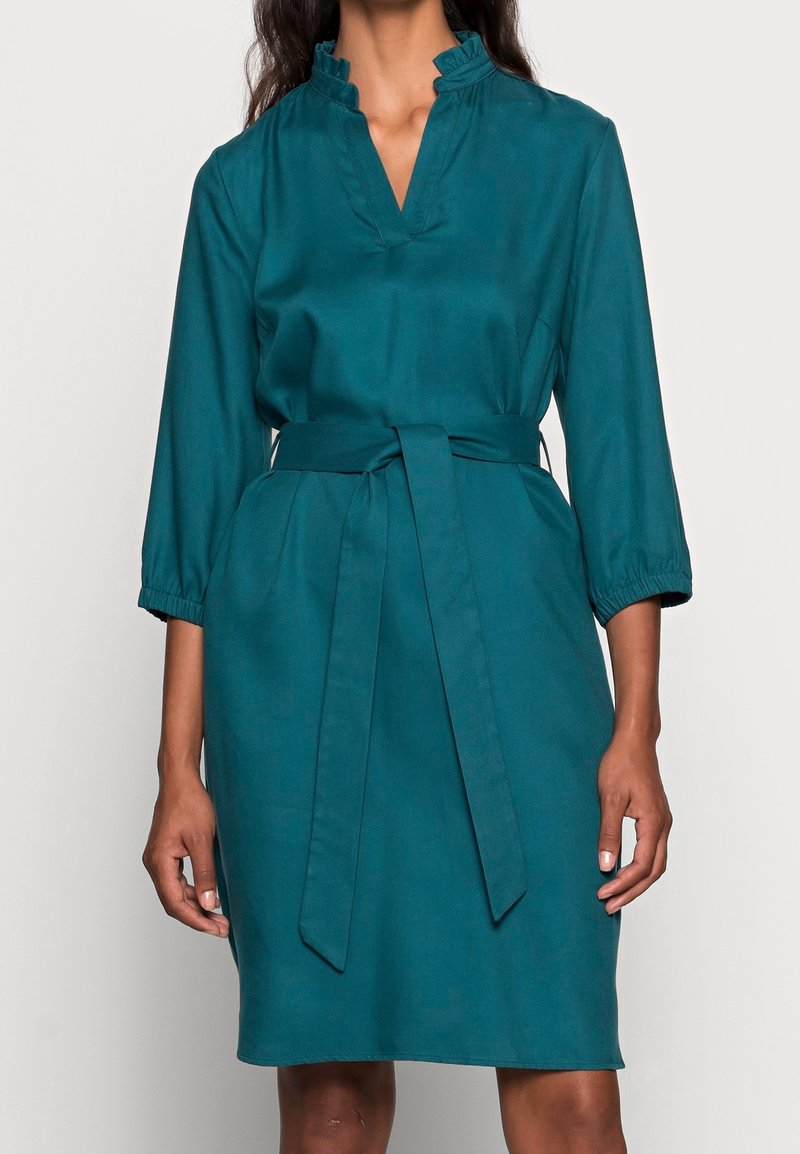 Teglrød kjole lavet af blødt stof med flæsekant ved kraven, 3/4-ærmer og et knyttet bælte i taljen for en markeret silhuet.