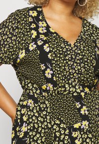Schwarzes Blumenkleid mit V-Ausschnitt, kurzen Ärmeln und verschiedenen gelben und lila Blumenmustern sowie Knopfakzenten. Texturierter Stoff.