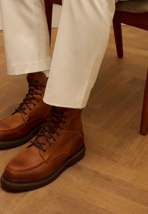 Bruine leren enkelboots met donkere veters, rubberen zolen en stikdetails; gedragen met crèmekleurige broeken op een houten vloer.