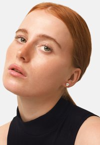 Orecchini a bottone tondi con pietra trasparente incastonata in metallo color argento, indossati da un modello con capelli rossi, vestito con un dolcevita nero a righe.
