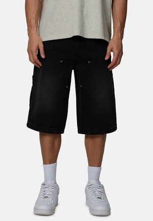 Mężczyzna ubrany w czarne cargo do kolan, jasnoszary t-shirt, białe skarpetki sięgające za kostkę oraz białe trampki, stoi z rękami swobodnie opuszczonymi wzdłuż ciała.