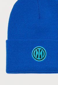 Μπλε πλεκτό σκουφάκι με αναδιπλούμενη περιφέρεια που διαθέτει έναν κυκλικό, τυρκουάζ λογότυπο σε μαύρο φόντο. Σχεδίαση υφής υφάσματος.