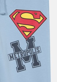 Gaiši zili sporta bikses ar lielu sarkanu un dzeltenu Supermena logo un tumši zilu tekstu "METROPOLIS" ar stilizētu "M". Mīksta auduma ar gludu tekstūru.