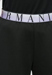 Pantaloni de trening negri cu o bandă elastică lată, de culoare deschisă, pe care este inscripționat textul "ARMANI" cu litere negre îndrăznețe. Textură netedă, design casual.