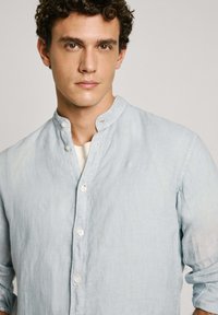 Svetlo modra lanena srajca z mandarin ovratnikom, belimi gumbi in diskretno izvezenim logotipom na prsih. Gladka tekstura, sproščen dizajn.