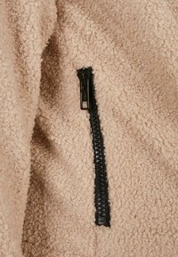 Beiges Fleece-Material mit einer strukturierten, lockigen Oberfläche; besitzt einen schwarzen Reißverschluss mit einem markanten Zipper auf der rechten Seite.