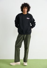 Felpa nera con logo rosa sul petto, abbinata a pantaloni a gamba larga verde oliva e sneaker beige con dettagli rossi. Sfondo semplice.