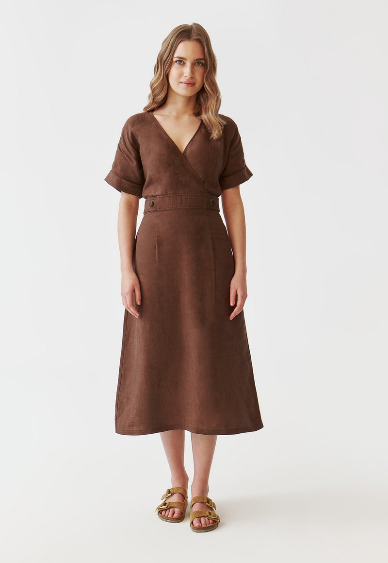 Braunes Leinenkleid mit Wickeldesign, kurzen Ärmeln, Seitentaschen und Knopfdetails. Kombiniert mit gelben Sandalen.
