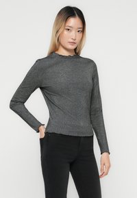 Młoda kobieta z prostymi blond włosami, ubrana w dopasowaną, długos sleeve, ciemnoszary prążkowany top i czarne spodnie, stojąca na jednolitym tle.