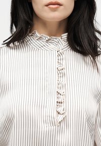 Γυναίκα που φοράει λευκό πουκάμισο με λεπτές κάθετες μαύρες ρίγες, με βολάν στον ψηλό γιακά και βολάν στη λωρίδα με κουμπιά.