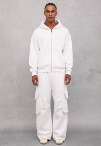 Sudadera blanca con cremallera y bolsillos tipo canguro, combinada con pantalones cargo de corte holgado que cuentan con bolsillos laterales y puños acanalados. Textura de tela casual en toda la prenda.