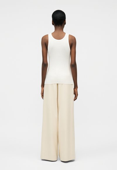 Γυναίκα που στέκεται πλάτη, φορόντας ένα λευκό ριμπ τοπ χωρίς μανίκια και φαρδιά κρεμ παντελόνια, σε απλό λευκό φόντο.