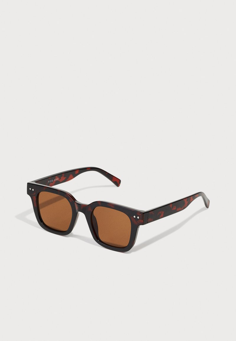 Lunettes de soleil en écaille de tortue avec une monture carrée épaisse, des verres marron et de subtils accents argentés sur les branches. Texture en plastique lisse.