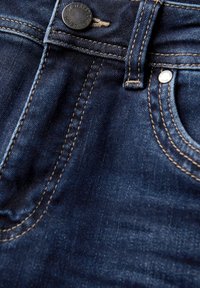 Jean en denim bleu foncé avec des coutures visibles, un bouton en métal, et une poche à pièces. Le tissu présente une surface légèrement texturée.