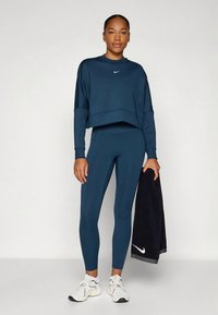 Tumszils cropped sporta džemperis ar apaļu apkakli un logotipu; atbilstošas legingi; grip apavi; turot tumšu dvieli. Gluda tekstūra, sportisks siluets.