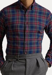 Koszula w kratę w kolorze granatowym, czerwonym i zielonym, z kołnierzykiem zapinanym na guziki i guziki z materiału. Noszona z dopasowanymi szarymi spodniami z bocznymi zaszewkami.