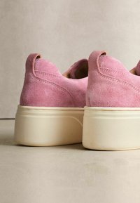 Różowe zamszowe buty wsuwane z okrągłym noskiem, teksturowaną cholewką i masywną białą gumową podeszwą. Posiadają subtelną pętelkę z tyłu.