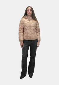 Giacca imbottita beige con zip frontale, design trapuntato, polsini elastici e cappuccio, indossata sopra jeans scuri. Ambientata su uno sfondo bianco.