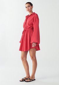 Rotes Hemdkleid aus leichtem Stoff mit lockerer Passform, elastischem Taillenbund, langen Ärmeln und ausgestelltem Saum. Kombiniert mit schwarzen flachen Sandalen.