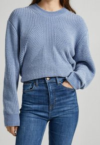 Suéter de punto azul claro con un patrón texturizado y puños acanalados, combinado con jeans oscuros de cintura alta. Diseño simple y casual.