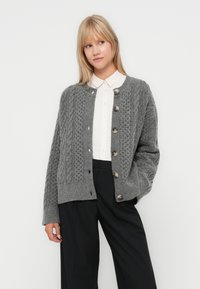 Grijze gebreide cardigan met een getextureerd kabelpatroon, voorzien van grote knopen en een ontspannen pasvorm. Gemaakt over een overhemd met kraag en een broek.