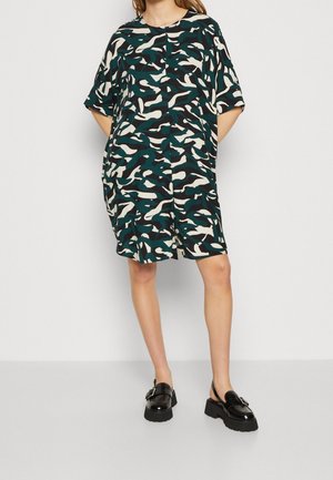 Vrouw draagt een loszittende jurk tot op de knie met een groen, zwart en wit abstract camouflagepatroon en zwarte loafers met dikke hak.
