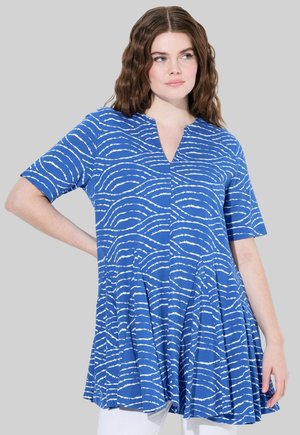 Túnica corta de manga corta azul con escote en V y patrón de olas blancas, que cuenta con un dobladillo acampanado y tejido suave y texturizado. Combinada con pantalones blancos.
