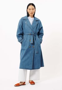 Denim trenchcoat met een getailleerde taille, brede revers en dubbelzijdige knopen, gecombineerd met witte wijde broek en oranje slides.