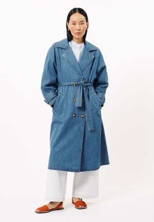 Denim trenchcoat met een getailleerde taille, brede revers en dubbelzijdige knopen, gecombineerd met witte wijde broek en oranje slides.