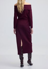 Robe bordeaux à épaules dénudées avec manches longues, silhouette ajustée et fente latérale, confectionnée dans un tissu lisse, associée à des bottes en cuir brillantes jusqu'aux genoux.