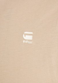 Béžová bavlnená tkanina s textúrovaným povrchom a bielym vyrazeným logom "RAW". Jednoduchý, moderný dizajn bez ďal�ších vzorov alebo akcentov.